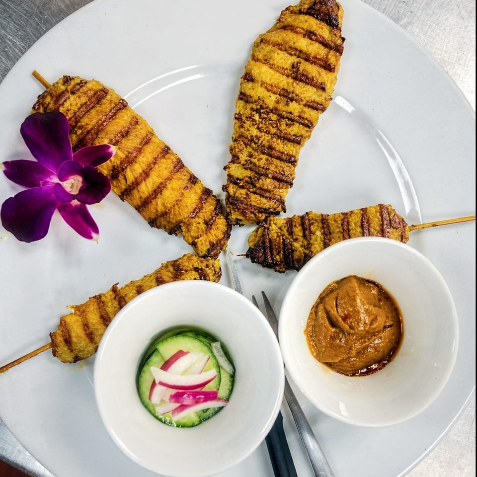Grilled chicken satay skewers with peanut sauce, cucumber salad, and a purple orchid on a white plate.